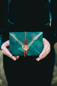 Elegant teal gift box adorned with golden accents and a leather tie, held delicately in a hand.