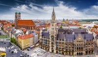 A panoramic view of Munich's historic town square showcasing iconic architecture, including the New Town Hall and the twin towers of St. Peter's Church under a dramatic sky.