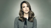 Jessica Alba en un elegante suéter gris con suaves ondas en su cabello, posó contra un fondo apagado.