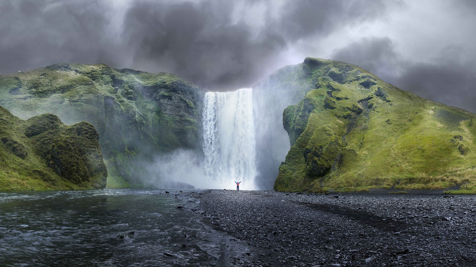 Una persona de pie frente a una cascada con un cielo nublado (manzana, cuerpo de agua, naturaleza, cascada, recursos hídricos)