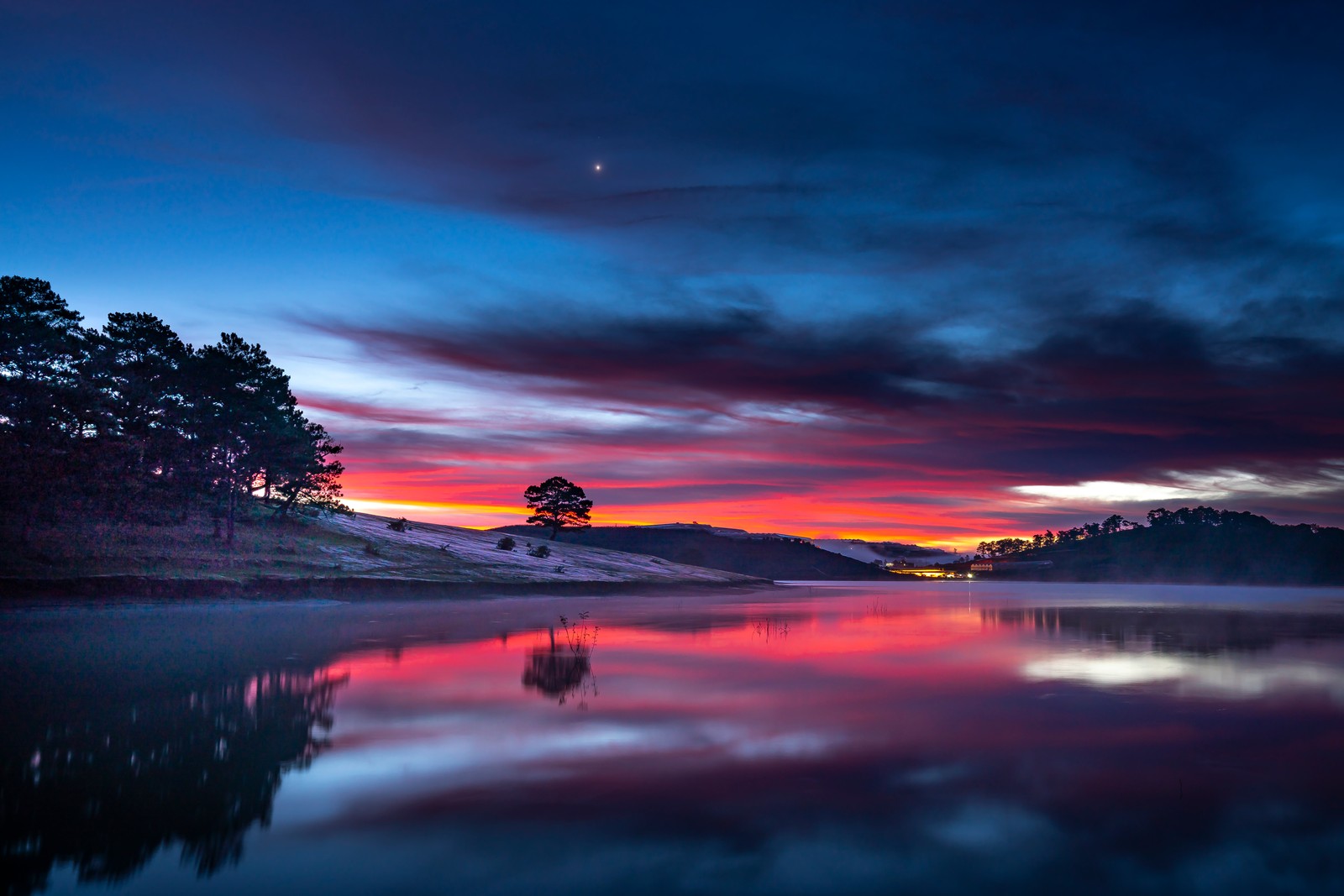 Una vista de un lago con un árbol y una luna en el cielo (atardecer, hora dorada, lago, cuerpo de agua, cielo nublado)