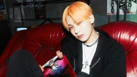 Sunoo of ENHYPEN poses on a vintage red couch, holding a skateboard, with a stylish streetwear look and striking orange hair.