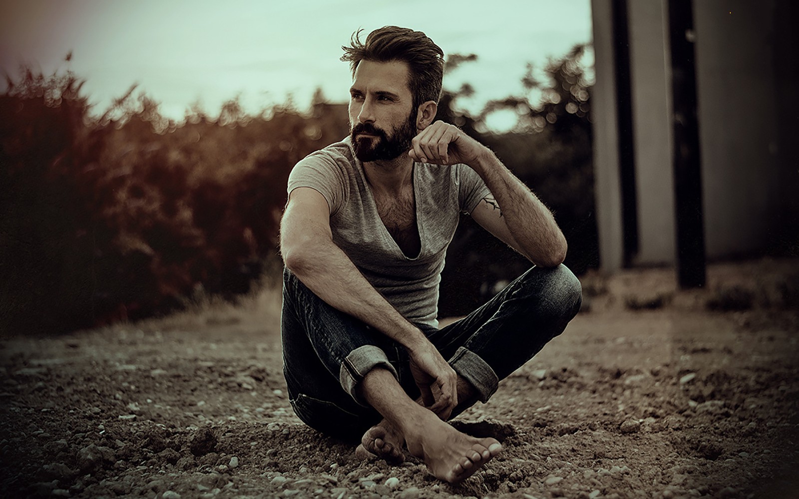 There is a man sitting on the ground with his hand on his chin (portrait, facial hair, sitting, human, portrait photography)