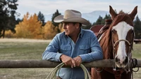 A cowboy in a blue shirt and hat leans against a wooden fence, gazing thoughtfully into the distance, with a horse by his side and autumn foliage in the background.