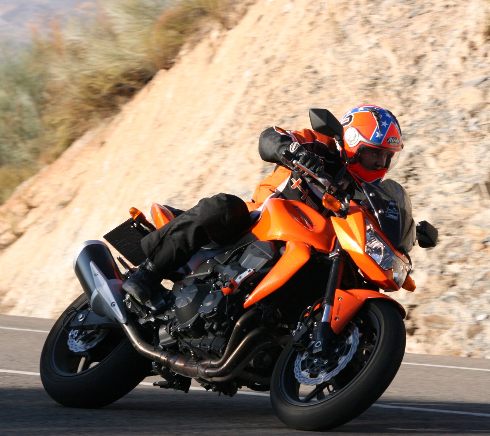 There is a man riding a motorcycle on a road near a hill (kawasaki z750, moto, naranja)