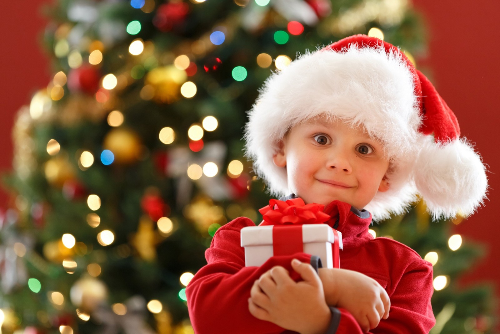 Criança fantasiada de papai noel segurando um presente em frente a uma árvore de natal (presente, feriado, natal, papai noel, vermelho)