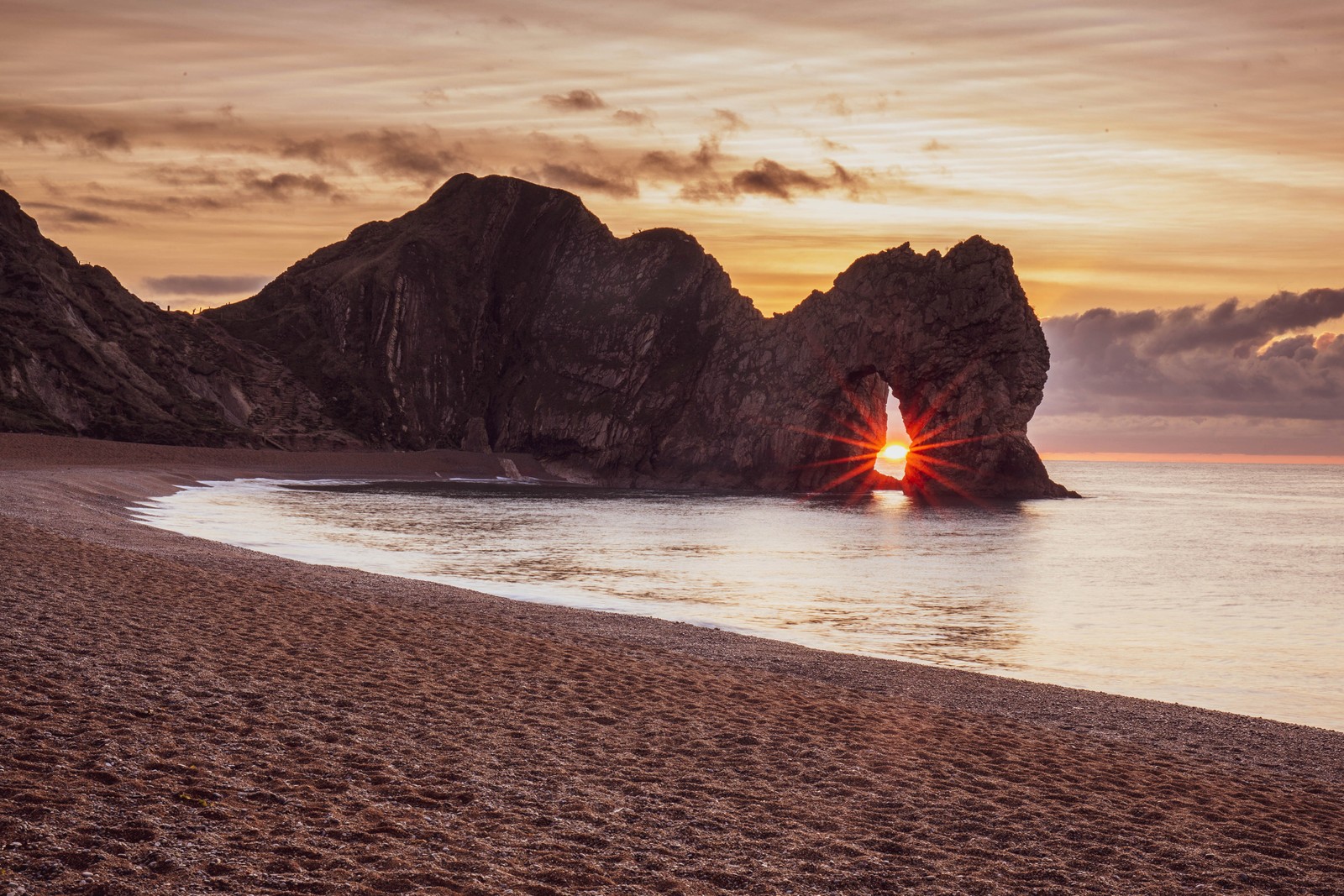 Uma formação rochosa na praia com um pôr do sol ao fundo (costa, por do sol, nascer do sol, natureza, mar)