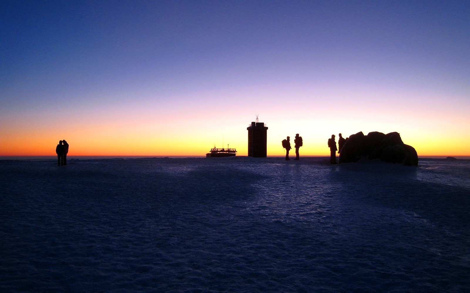 Pessoas em pé na praia ao pôr do sol com um farol ao fundo (horizonte, mar, por do sol, oceano, nascer do sol)