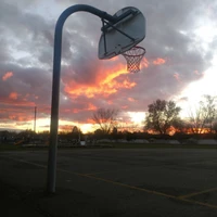 Pôr do sol sobre uma quadra de basquete vazia com uma cesta silhuetada contra nuvens coloridas.