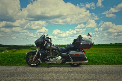 Harley Davidson Fat Boy garé sur une route pittoresque sous un ciel nuageux, entouré de champs verdoyants.