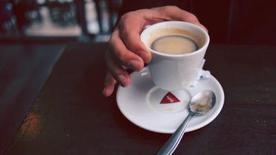 Uma mão segurando uma pequena xícara de café cheia de espresso, descansando em um pires com uma colher ao lado, sobre uma mesa de madeira escura.