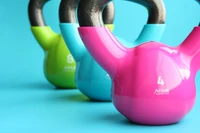 Kettlebells coloridos em tons vibrantes de rosa, azul e verde, dispostos contra um fundo azul brilhante, exibindo equipamentos de exercício para treinamento de força e peso.