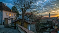 Crepúsculo sobre una ciudad checa, con una granja histórica y un árbol, con el río Vltava y una torre distante bajo un cielo dramático.