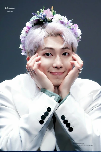 RM do BTS usando uma coroa de flores, sorrindo calorosamente enquanto descansa o queixo nas mãos.