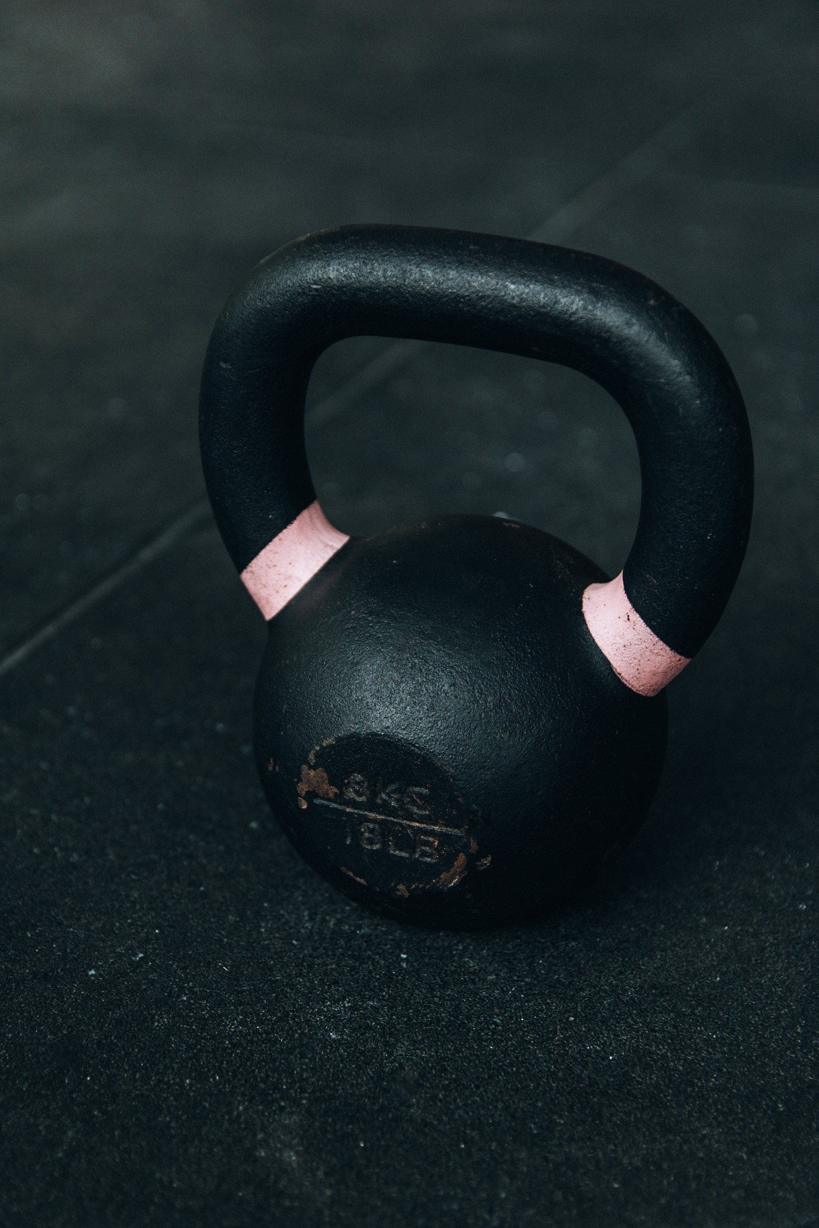 There is a black kettlebell with a pink handle on a black floor (kettlebell, exercise, dumbbell, fitness centre, weights)