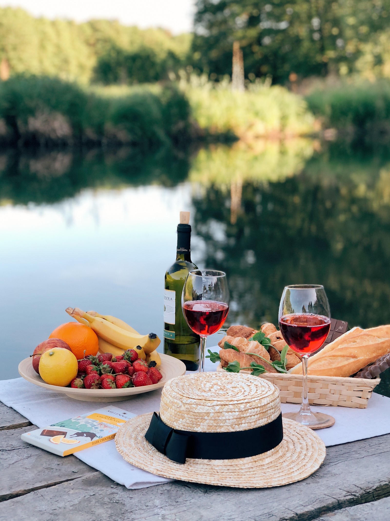 Hay un sombrero, un sombrero de paja, frutas y pan en una mesa de picnic (copa de vino, desayuno, vino, brunch, cristalería)