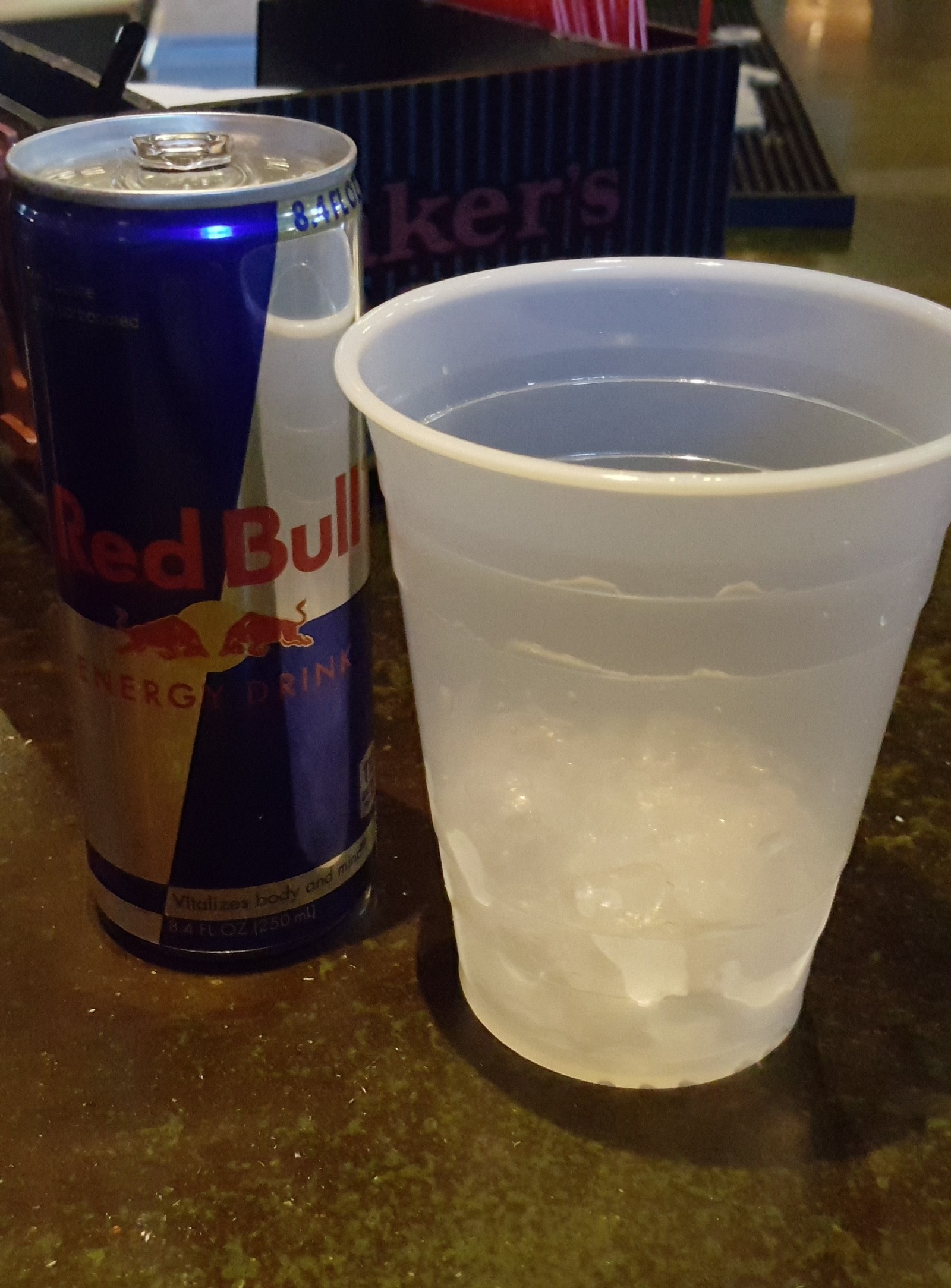 There is a plastic cup and a can of red bull on a table (alcohol, bar, bull, club, drink)