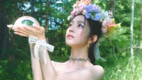 Irene in a floral crown, holding a crystal orb in a lush, green forest setting.