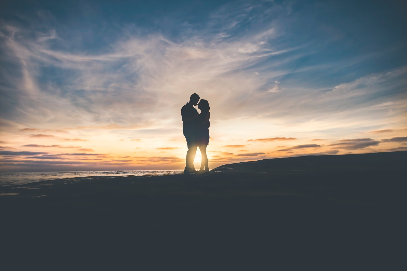 Um casal em pé na praia ao pôr do sol com os braços em volta um do outro (casal, pessoas na natureza, nuvem, horizonte, por do sol)