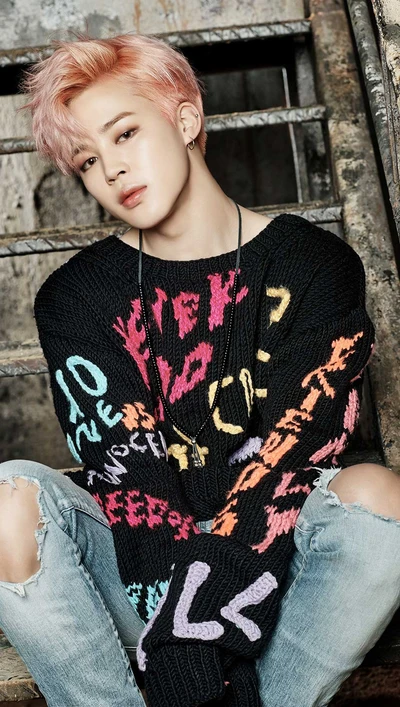 Jimin do BTS em um suéter oversized colorido, sentado contra um fundo industrial com uma expressão contemplativa.