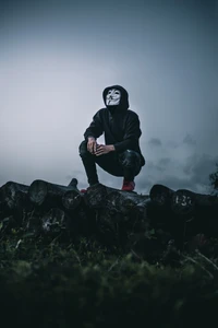 Figure in a Guy Fawkes Mask Sitting on Logs Amidst Misty Mountains