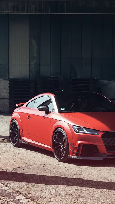 Элегантный красный Audi TT RS припаркован в промышленной обстановке, демонстрируя свой спортивный дизайн и агрессивную стойку с черными дисками и эффектной решеткой.