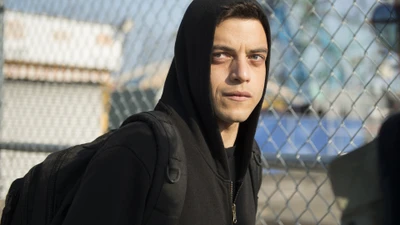 Rami Malek como Mr. Robot na moda de rua, vestindo um moletom com capuz e uma mochila contra um fundo de cerca de arame.