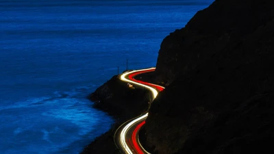 Route côtière nocturne illuminée par les phares des voitures contre un océan bleu