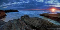 Amanhecer na Praia Whitehaven: As ondas quebram contra a costa rochosa sob um céu colorido.