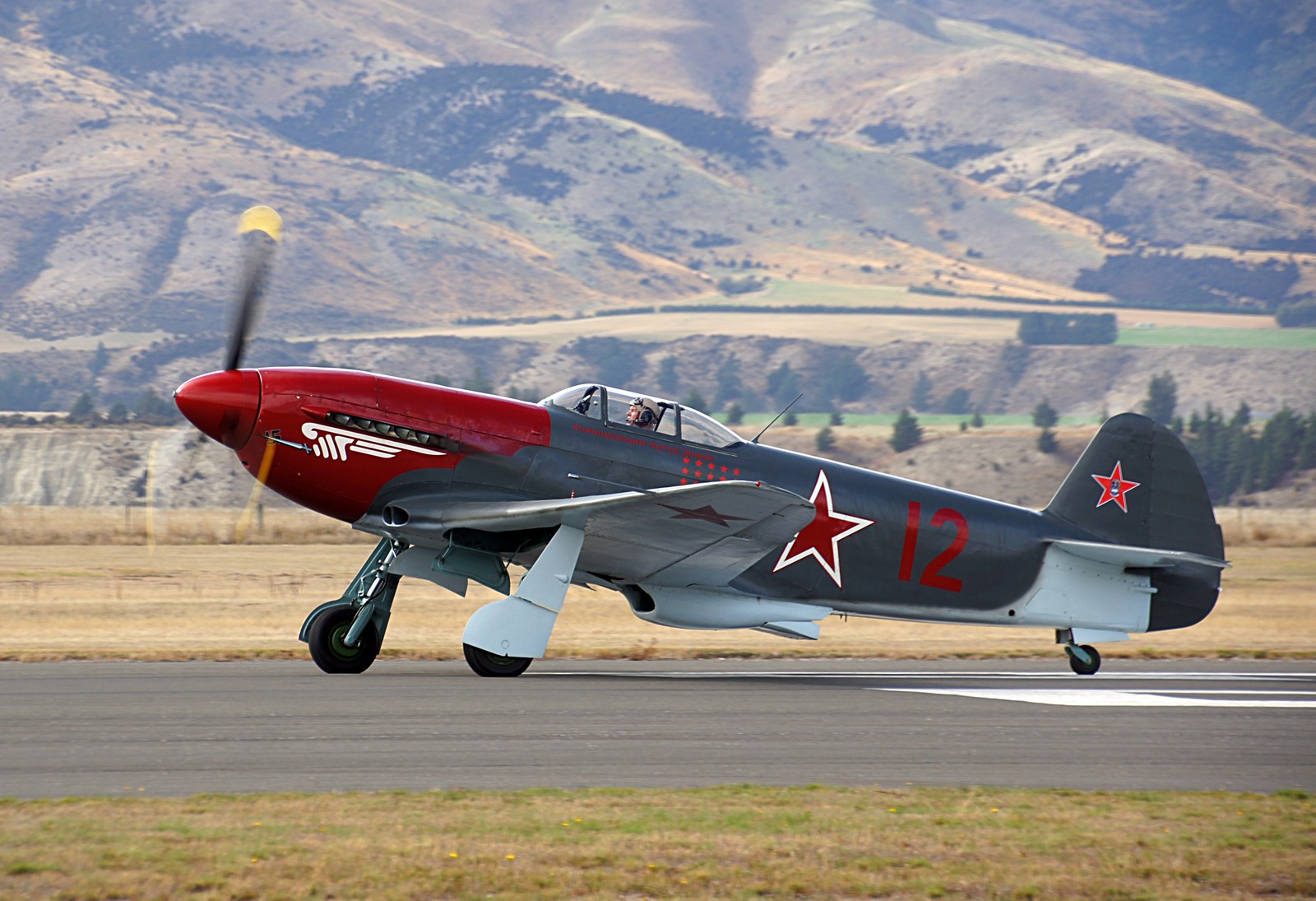 Hay un pequeño avión rojo y gris en la pista (yakovlev yak 1, avión militar, aeronave, avión, aviación)