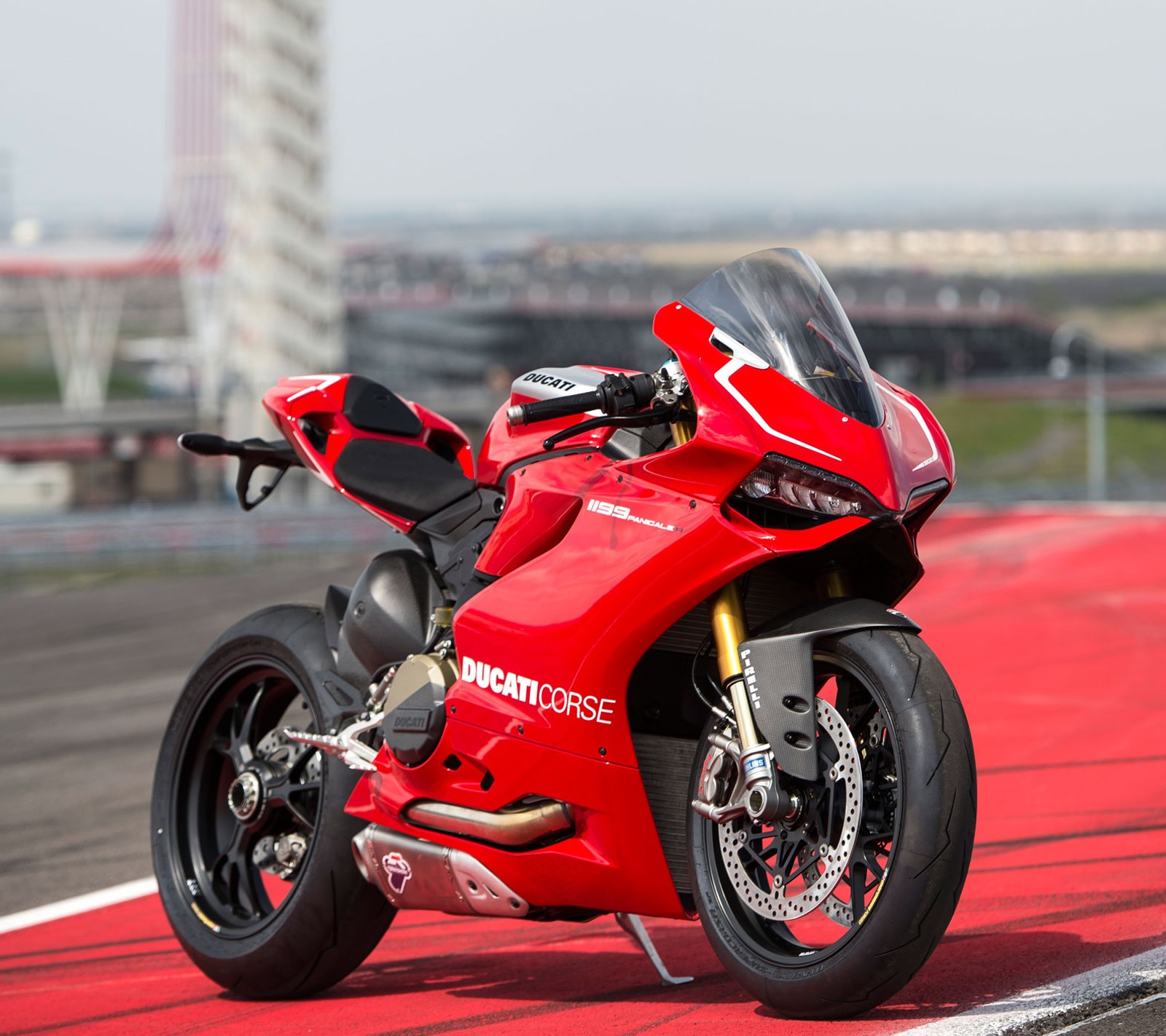 Une moto rouge garée sur une piste avec une ville en arrière-plan (1199, ducati, superbike)