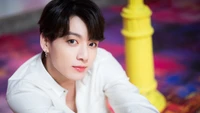 Jungkook en una elegante camisa blanca, posando con un fondo vibrante y un pilar amarillo.