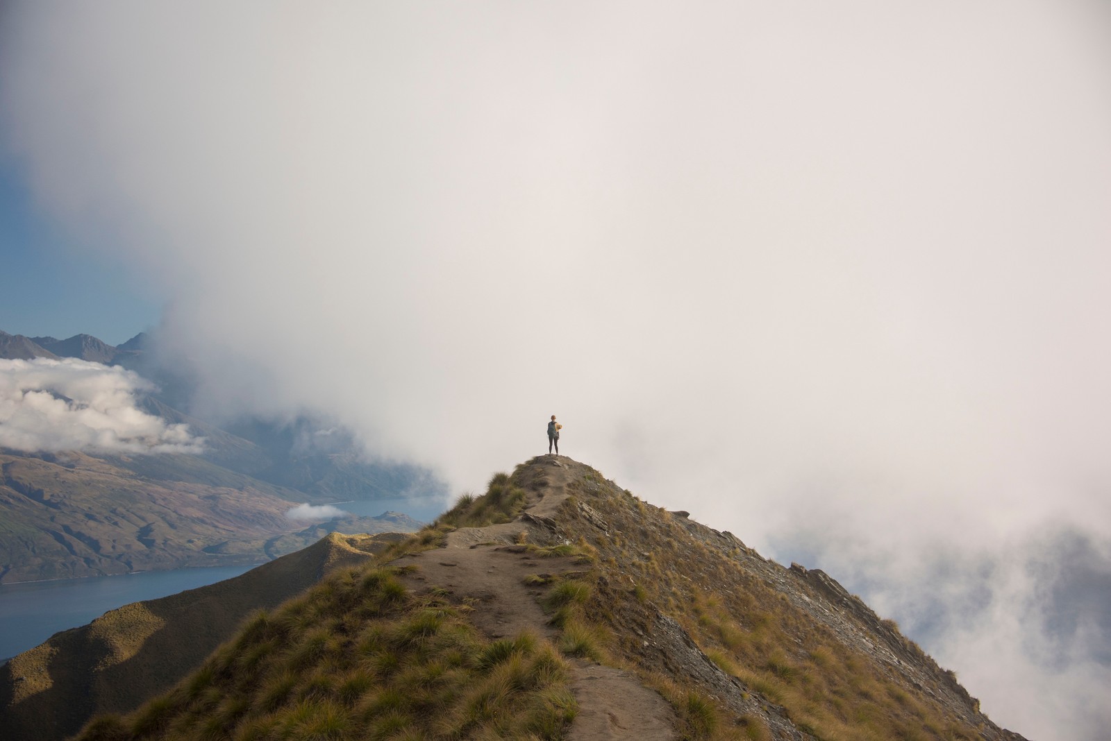 There is a man standing on a mountain top with a view of the water (mountainous landforms, mountain, highland, ridge, mountain range)