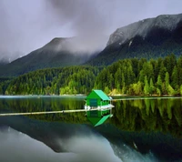 Casa verde serena en un lago tranquilo rodeado de montañas y nubes