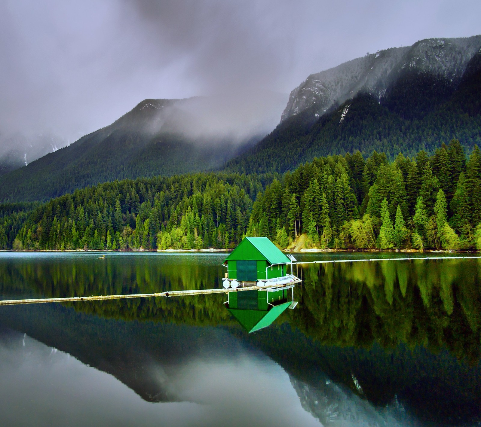 Il y a une petite maison verte sur un petit quai au milieu d'un lac. (nuages, maison, lac, montagne)