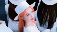 Vivi do LOONA com cabelos laranja marcantes, usando um chapéu branco, cercada por colegas em trajes elegantes.