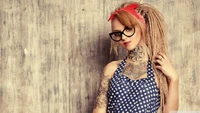 Modisches Mädchen mit einem Ärmeltattoo und stylischen Brillen, das eine verspielte Frisur und ein retro-inspiriertes Outfit vor einem strukturierten Hintergrund präsentiert.