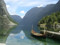 Paisagem de fiorde serena com um barco tradicional atracado em um lago tranquilo, emoldurado por montanhas majestosas e vegetação exuberante.