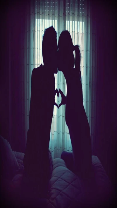 Silhouette of a couple forming a heart with their hands against a softly lit window.