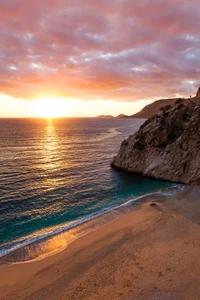 Pôr do sol sobre uma praia serena cercada por penhascos e ondas suavemente batendo na costa.