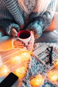 Scène matinale confortable avec une personne tenant une tasse de café, entourée de lumières chaudes et d'un livre sur une douce couverture.