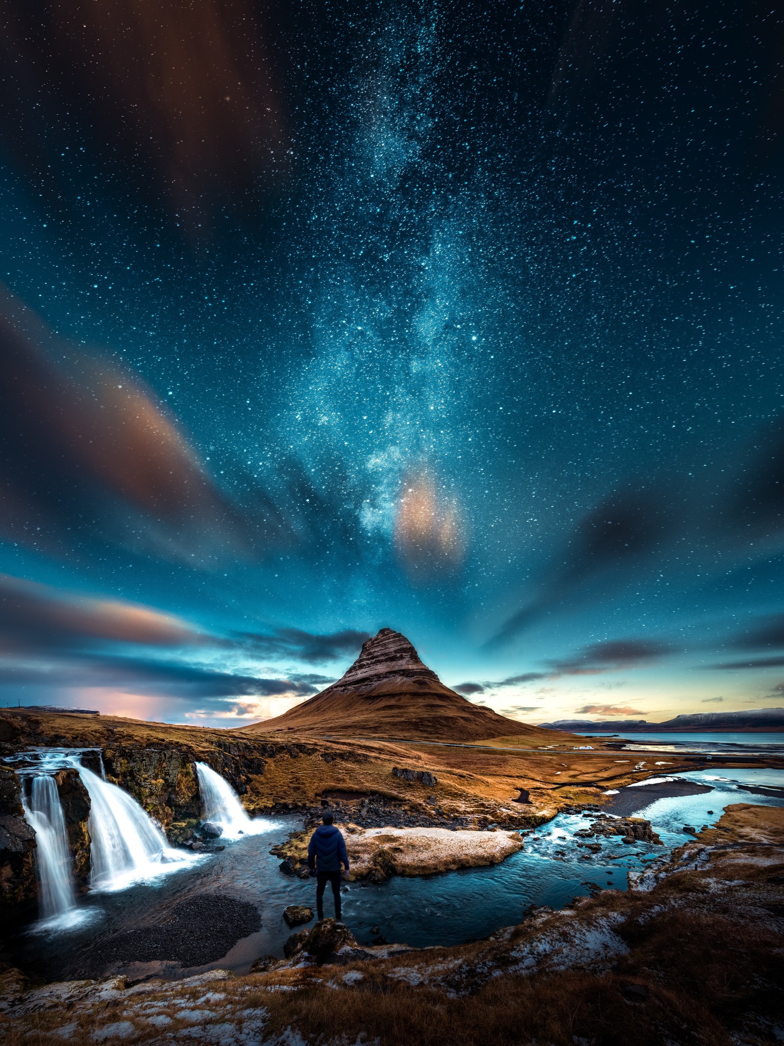 Un homme debout sur un rocher regardant une cascade sous un ciel étoilé (noir, montagne, nuit, ciel, étoiles)