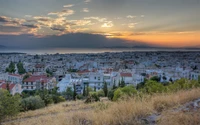 Pôr do sol sobre Atenas: uma vista panorâmica da cidade com telhados, árvores e o horizonte iluminando o céu da noite.