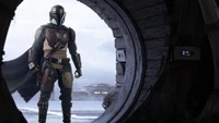 The Mandalorian stands poised at the entrance of a circular doorway, gazing out into a desolate landscape.