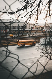 A yellow bus navigating a snowy, urban landscape, framed by bare tree branches and a chain-link fence.