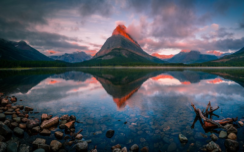 Вид на гору с озером и камнями на переднем плане (swiftcurrent lake, восход солнца, отражение, облачно, раннее утро)