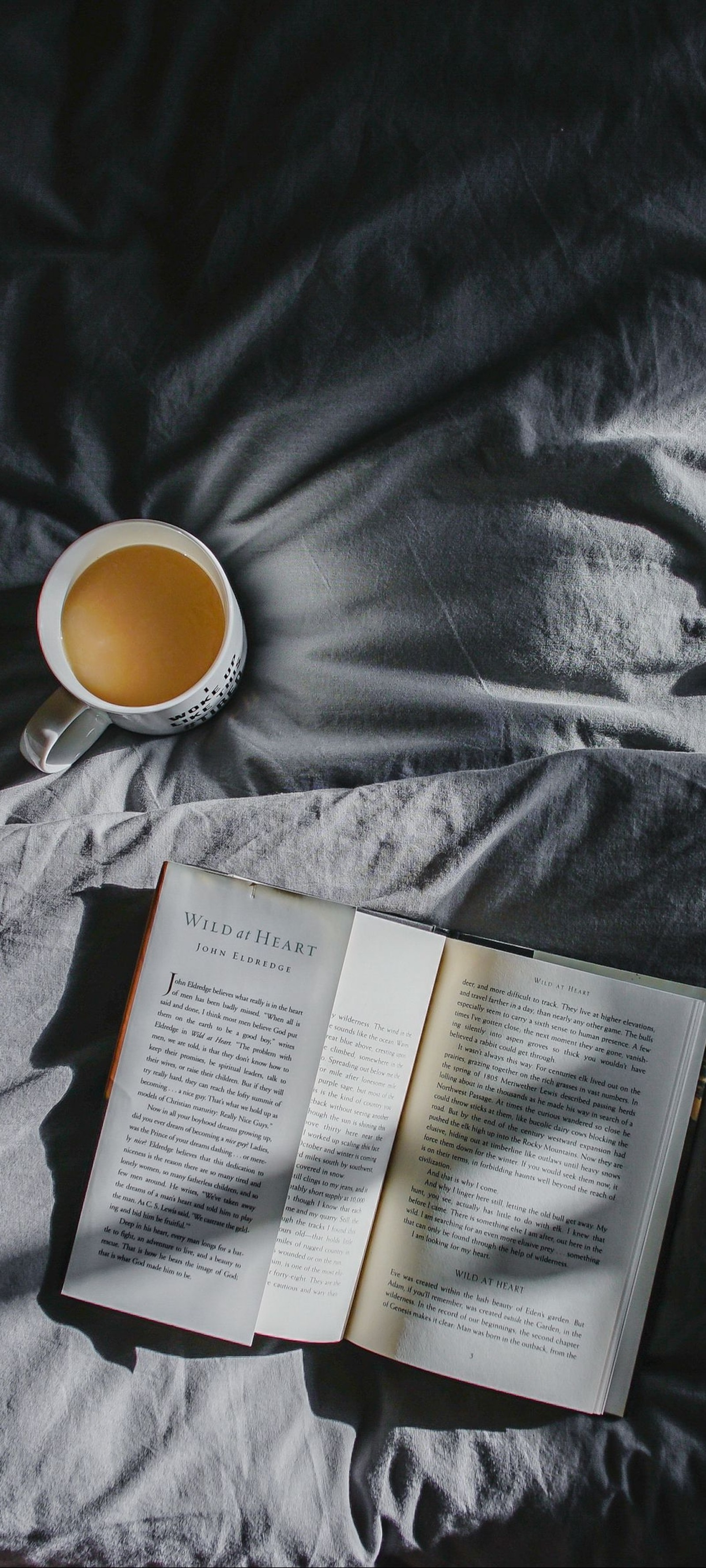 There is a book and a cup of coffee on a bed (book, coffee, tea, tableware, drinkware)