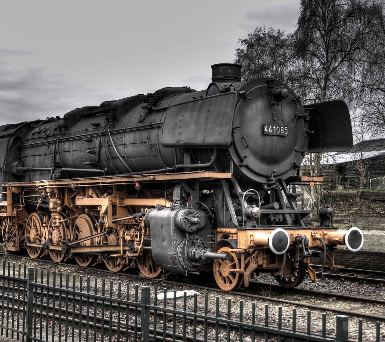 Un tren en las vías con una cerca y árboles al fondo (blanco y negro, agradable, ferrocarril, cielo, velocidad)