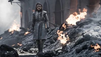 Arya Stark em meio à devastação da batalha, cercada por chamas e cinzas.