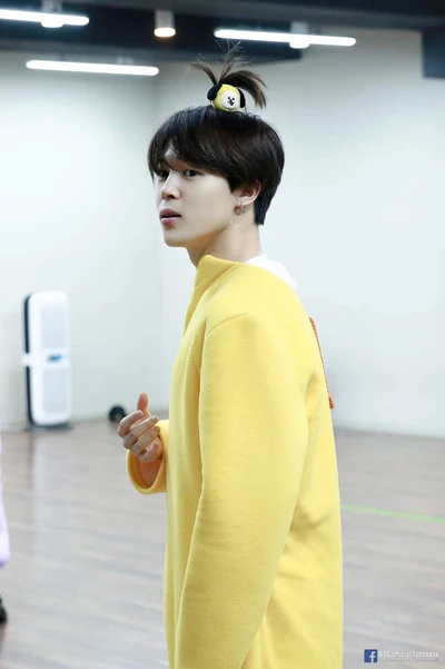 Jimin em um traje amarelo brincalhão com um penteado fofo, capturando o charme do K-pop.